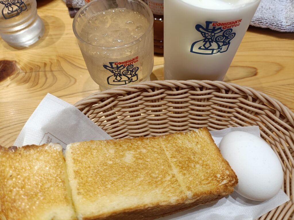 「コメダ珈琲店のモーニング。厚切りトーストとゆで卵が乗ったバスケットと、ロゴ入りカップのドリンクが木のテーブルに置かれている」