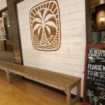 白い壁にヤシの木のロゴが描かれ、その前に木製ベンチが置かれたアロハテーブル店内の写真。