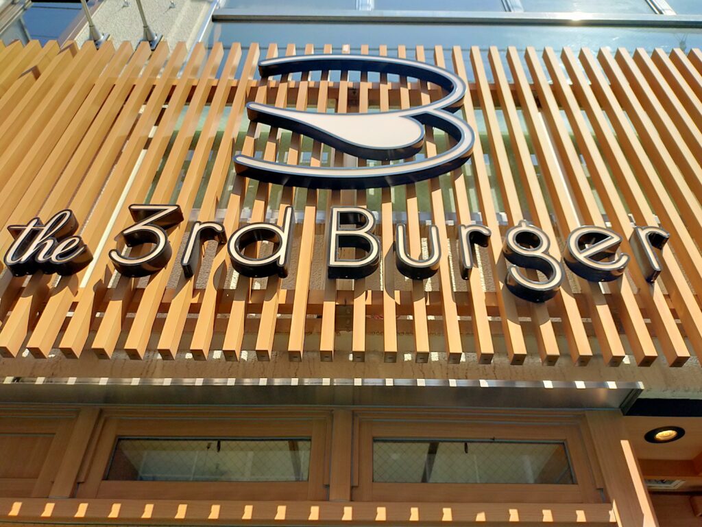 「木目調ルーバーの壁に白いロゴマークと『the 3rd Burger』の文字看板が付いた店舗外観の写真」