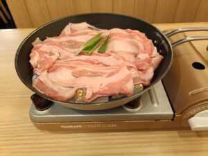 「火の音水の音 さいたま新都心店で提供されるすき焼き定食の鍋。カセットコンロの上のフライパンに豚肉とネギが生の状態で並んでいる」