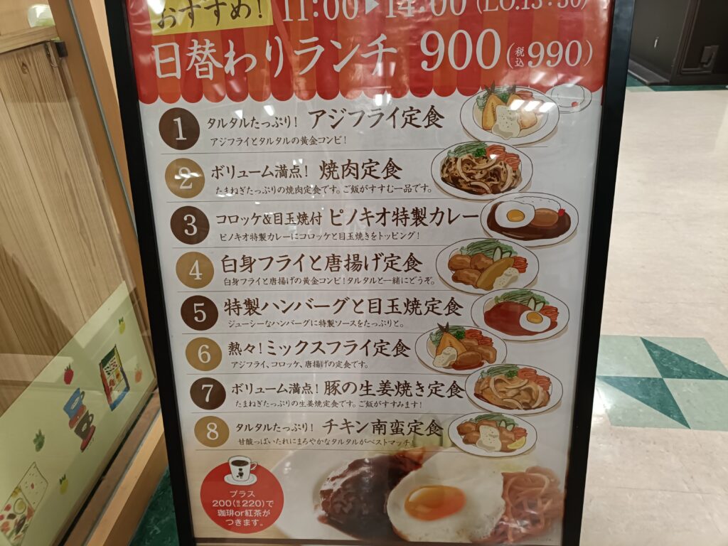 「喫茶店ピノキオの『日替わりランチ900円（税込990円）』と書かれたメニュー看板。曜日ごとの定食内容と料理写真が並んでいる」