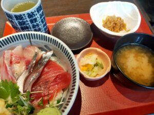 「刺身が盛られた海鮮丼と、小鉢や茶碗蒸し、味噌汁が赤いお盆に並んだまるがまるのランチセット」