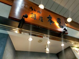 「天井からのライトに照らされた木製看板に『うどん 銀座 木屋』と書かれている店舗外観のアップ」