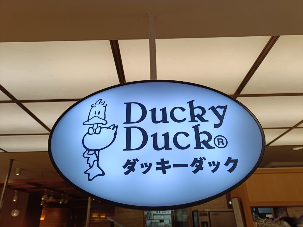 「天井から吊り下げられた丸いサインに、アヒルのイラストと『Ducky Duck ダッキーダック』の文字が描かれている店舗看板」