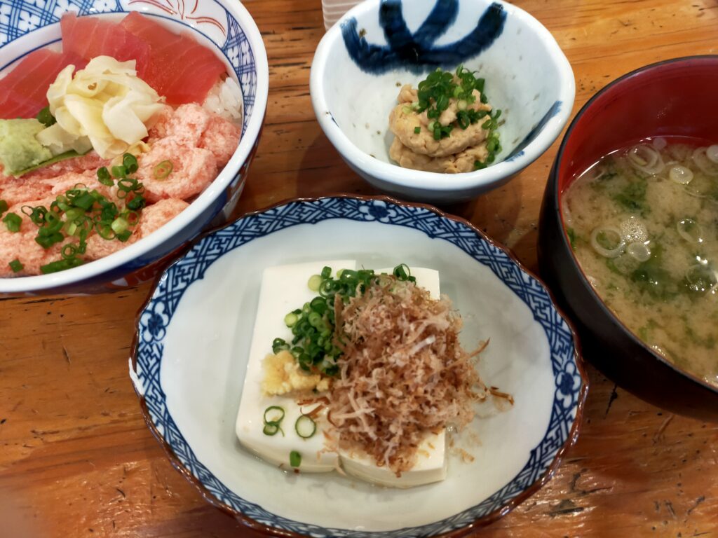 「かつお節と刻みネギがのった冷奴の小鉢を中心に、海鮮丼や味噌汁が並ぶ磯丸水産のランチセットのテーブル」