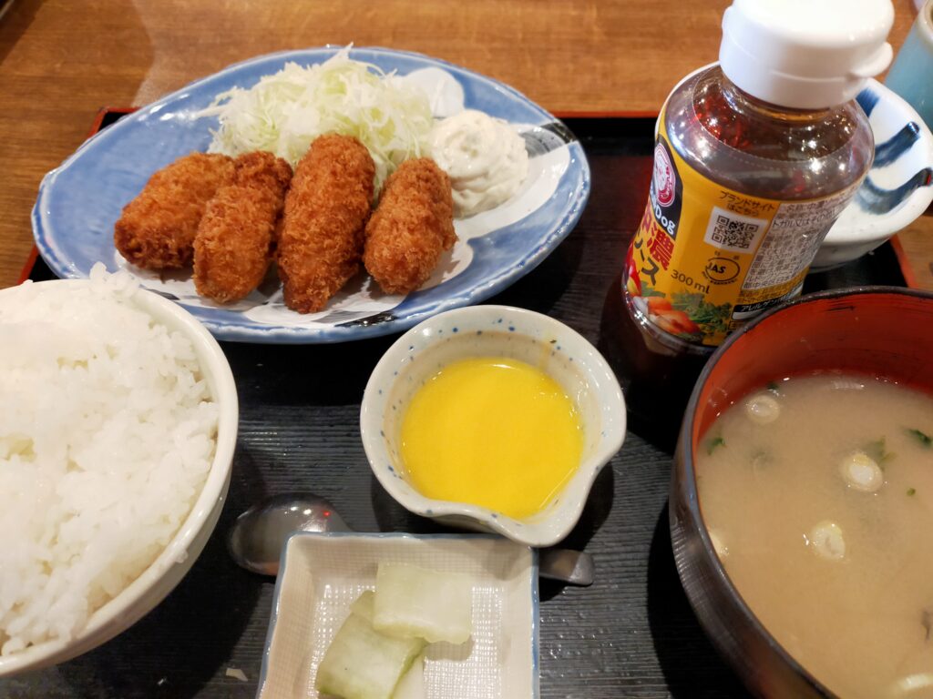 「皿に盛られた牡蠣フライとキャベツ、その周りにごはん、味噌汁、小鉢が並ぶ磯丸水産のフライ定食」