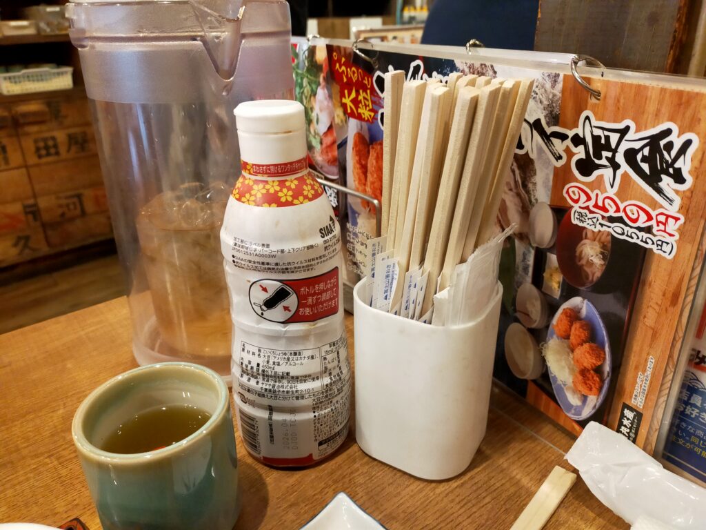 「磯丸水産のテーブルに置かれた調味料ボトル、割り箸立て、ランチメニュー冊子と湯のみ」