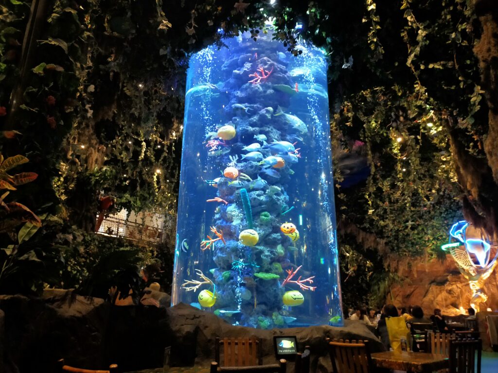 「青いライトで照らされた円柱型の大型水槽の中を、色とりどりの熱帯魚が泳ぎ、周囲にジャングル風の装飾がある店内風景」