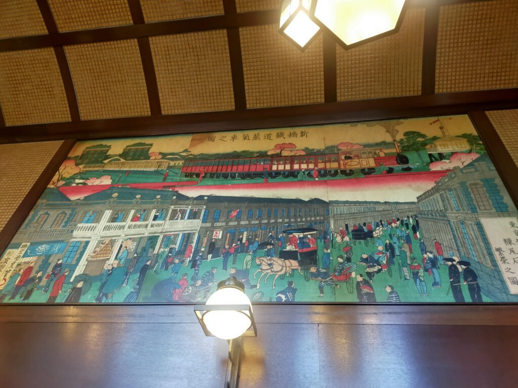 「木枠天井と照明の下にカラフルな街並みの絵画が掛けられた椿屋珈琲店内の壁面」