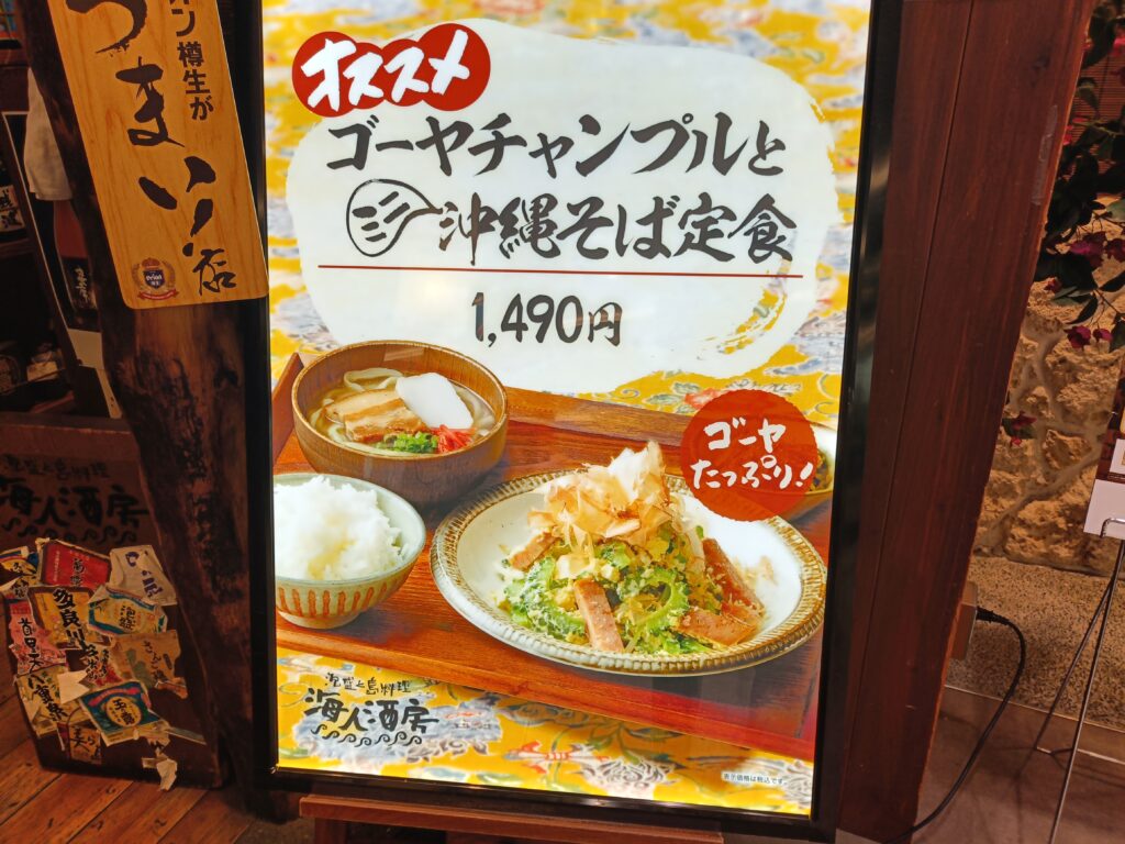 「ゴーヤチャンプルと沖縄そば定食を紹介する海人酒房のスタンドメニュー。料理写真と1490円などの価格が大きく表示されている」