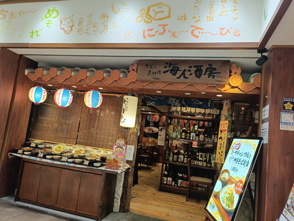 「海人酒房 池袋サンシャインシティ店の外観。木製カウンターの上に沖縄料理の食品サンプルが並び、奥にはボトル棚や提灯が見える店頭風景」