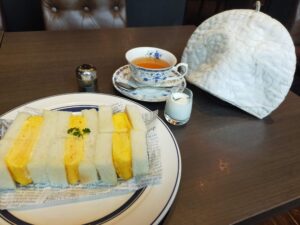 「厚焼き玉子を挟んだサンドイッチがのった大皿と、花柄のカップ＆ソーサーの紅茶、白いティーコジーとミルクピッチャーがテーブルに並んでいる」