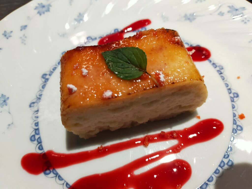 「長方形の焼き菓子風デザートの上に小さなミントの葉がのり、白い皿の上には赤いソースが線状にかけられているアップ写真」