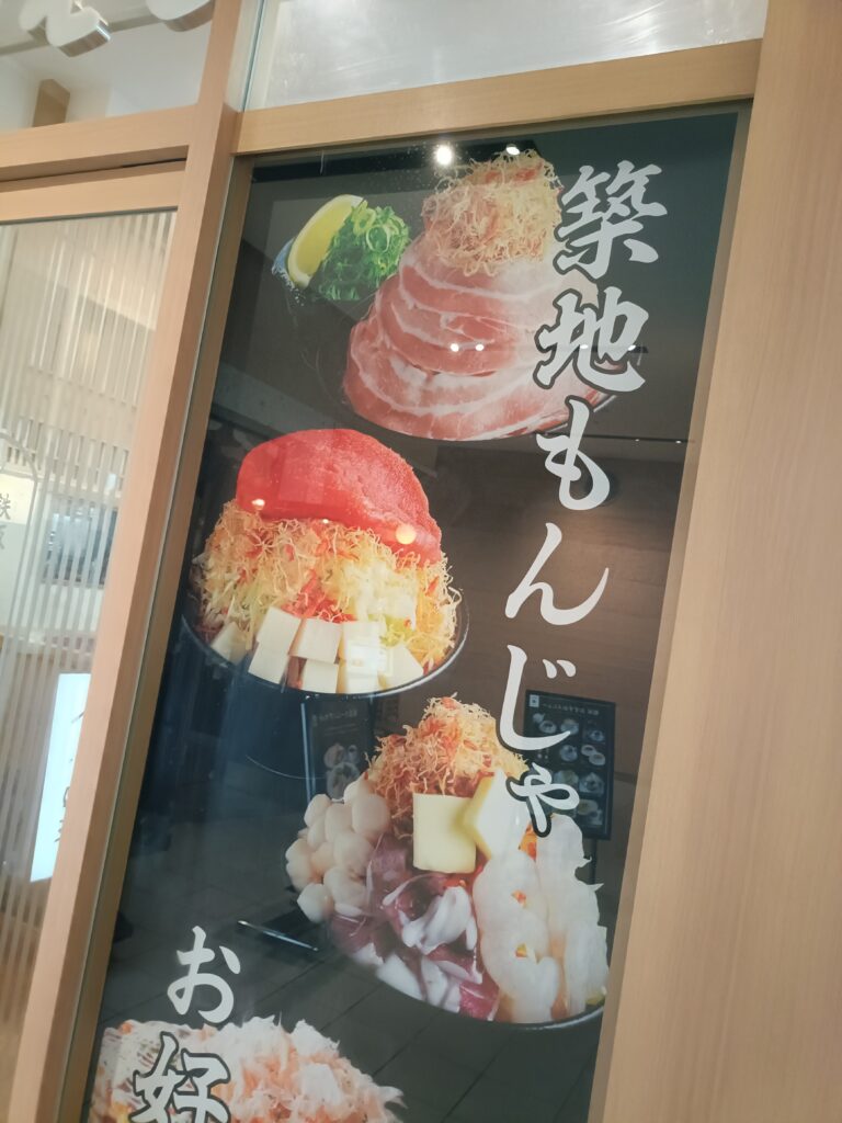 「『築地もんじゃ』の文字と、明太子やチーズ、シーフードなど具材が山盛りになったもんじゃ焼きの写真が3つ並んだこてがえし店頭ポスター」