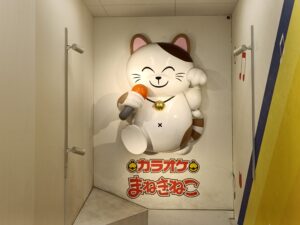 「白い壁にマイクを持った大きな招き猫の立体オブジェと『カラオケまねきねこ』のロゴが描かれた店内通路」