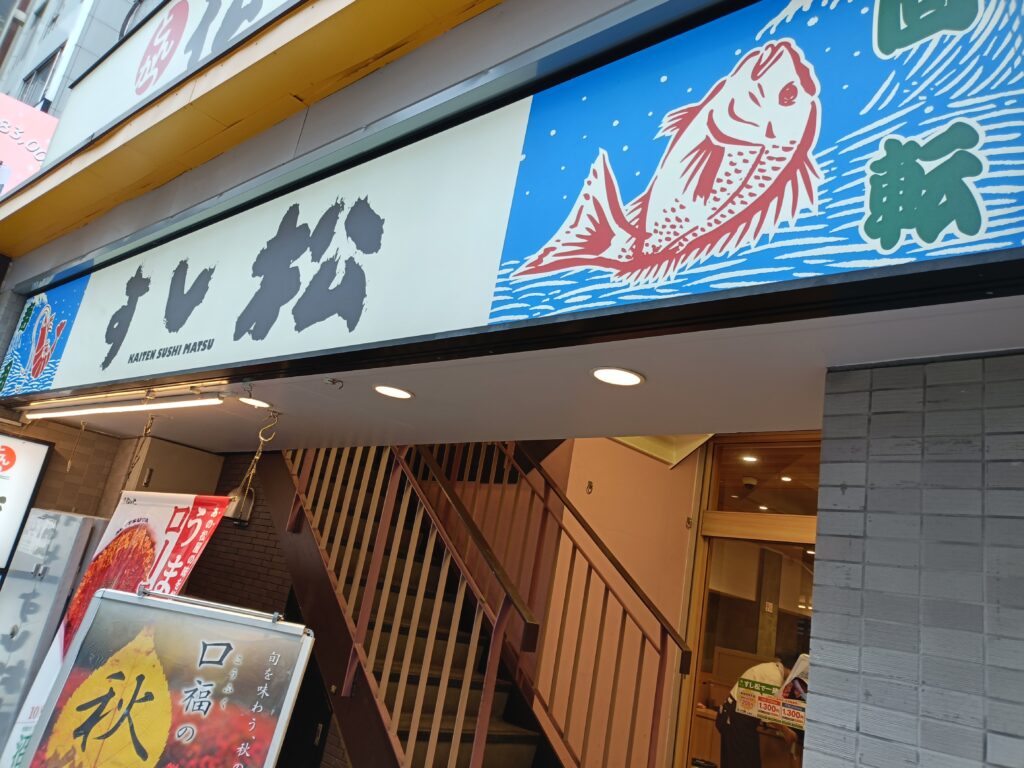 すし松の店舗入口付近の看板。店名と魚のイラストが見える外観。