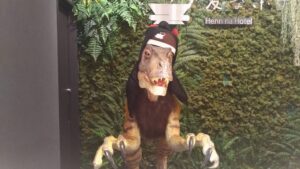 「変なホテル東京西葛西のフロントで、帽子をかぶった恐竜型ロボットがカウンター越しにお客様を出迎えている様子」