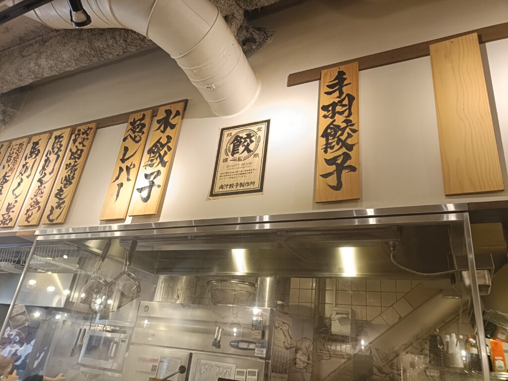 「肉汁餃子のダンダダンの店内カウンター越しに見える厨房と、頭上に吊り下がった『肉汁焼餃子』などの木札メニュー」