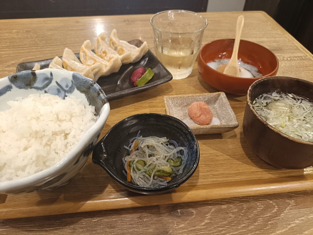 「木のトレーの上に焼き餃子の皿、ご飯茶碗、スープ、小鉢、漬物、ドリンクが並んだ肉汁餃子のダンダダンの餃子定食」