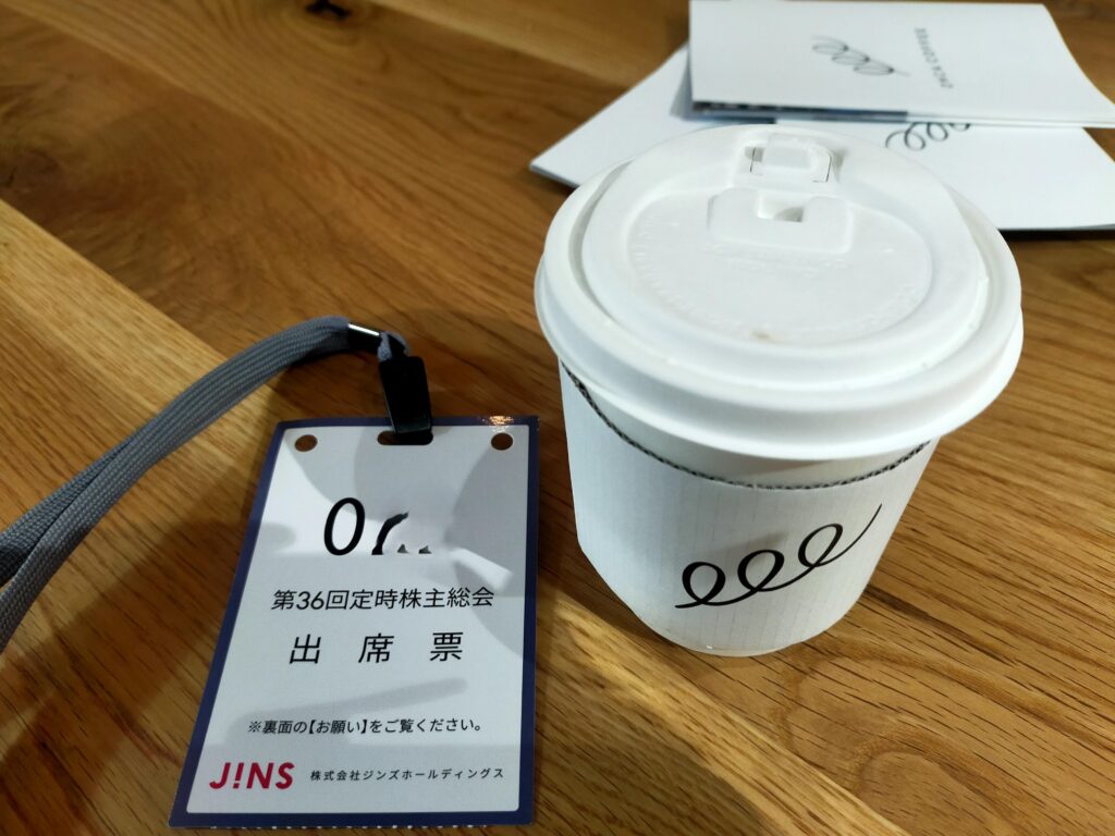 「JINS株主総会の会場テーブルに置かれたフタ付き紙コーヒーカップと、株主総会出席票のネームホルダー」
