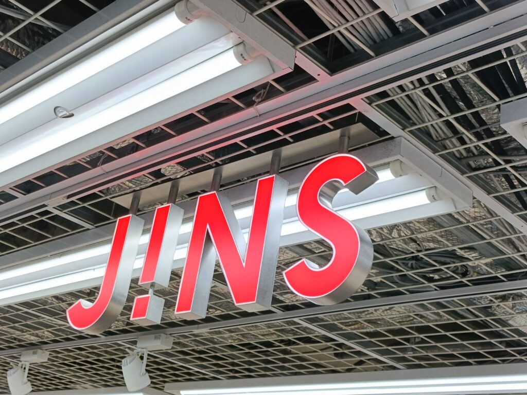 「天井の白い蛍光灯の下に赤いJ!NSロゴ看板が吊り下がった、ジンズ店舗の外観クローズアップ」