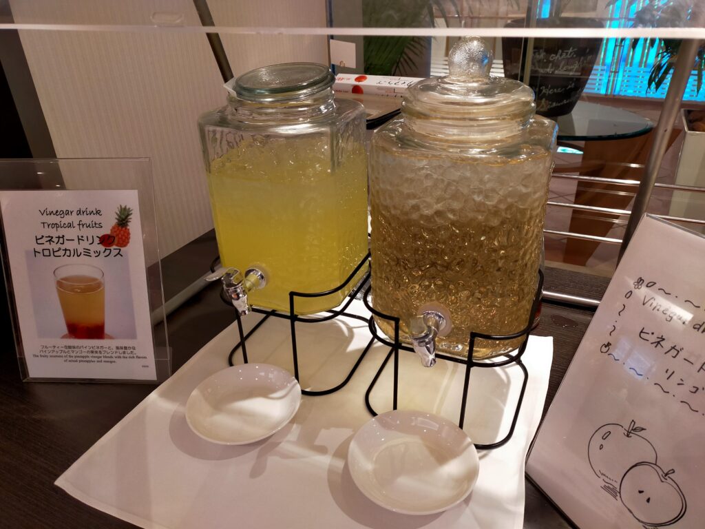 「スタンドに載せた2つの大型ガラスジャーにレモン入りウォーターなどが入っているビュッフェ形式のドリンクコーナー」