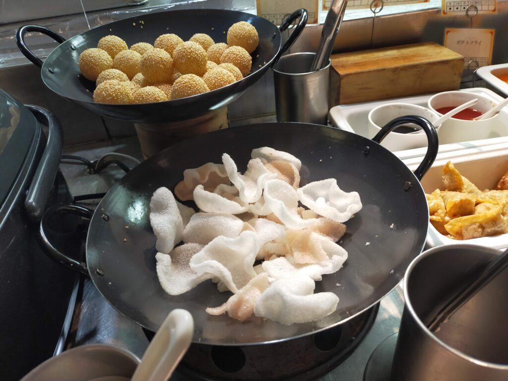 「丸いフライパンに白い点心風の生地が並び、その奥に丸い揚げ物が入った鍋が見える中華ビュッフェの調理コーナー」