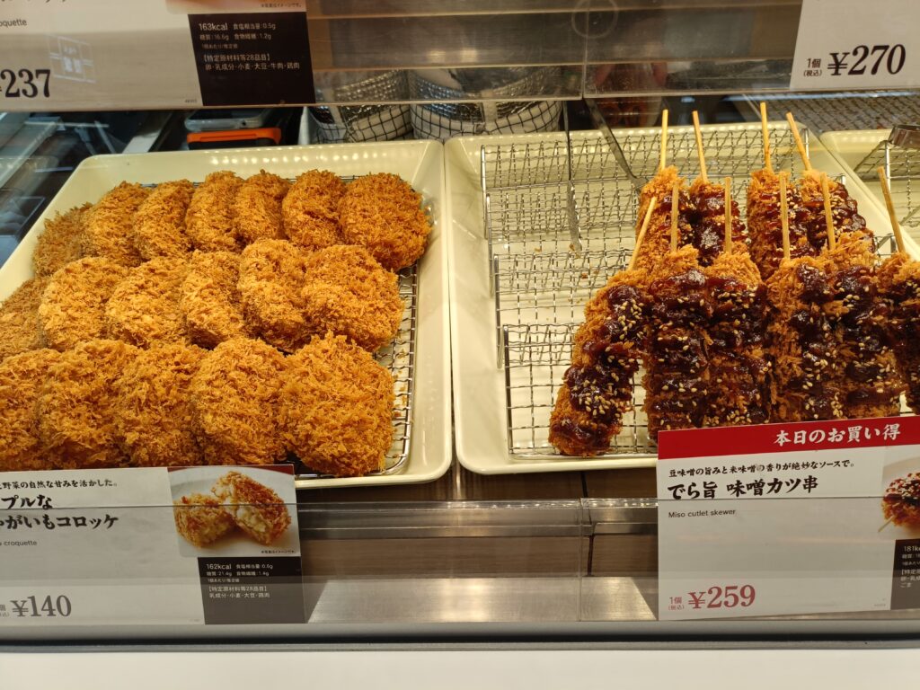 「ステンレスの網トレーの上に、円形のコロッケと細長いカツが整然と並んだ神戸コロッケのショーケース内」