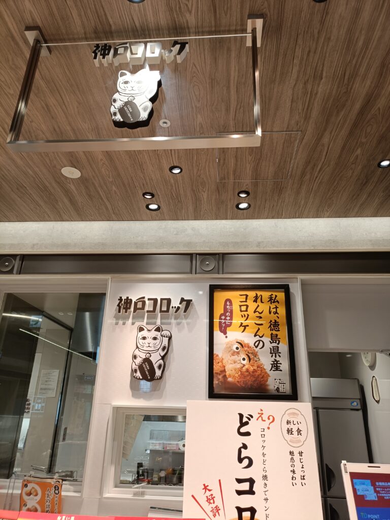 「木目の天井と照明の下に、神戸コロッケの案内ポスターやメニューが立てかけられている店頭カウンター」
