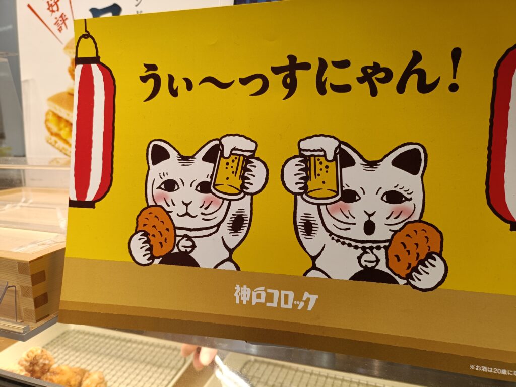 「黄色い背景に、ビールジョッキとコロッケを持った2匹の招き猫イラストとキャッチコピーが描かれた神戸コロッケの店頭ポップ」